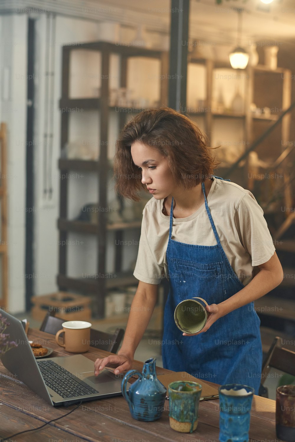 Craftswoman with handmade mug standing by table and looking at laptop screen while watching online video