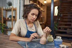 Young female artisan in workwear holding paintbrush and self-made jug while painting earthenware items in workshop