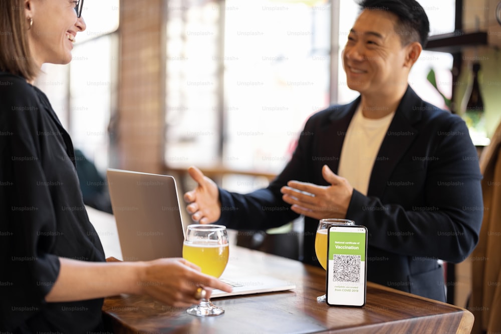 Multiracial friends drinks cocktails at table with smartphone with app of Coronavirus Vaccination in cafe. Concept of health protection during pandemic. Friendship. Happy caucasian woman and asian man