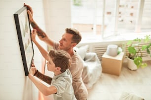 Retrato de alto ángulo de padre e hijo colgando cuadros en la pared mientras se mudan a un nuevo hogar, espacio de copia