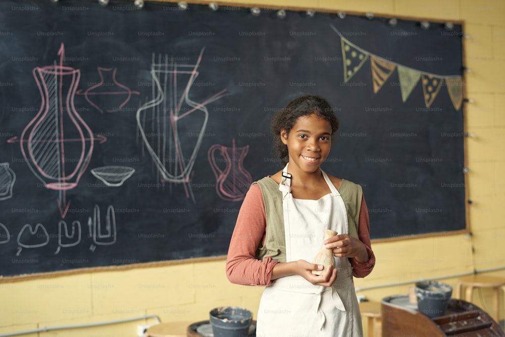 Happy cute schoolgirl with handmade clay pot standing by blackboard with sketches of earthenware in classroom