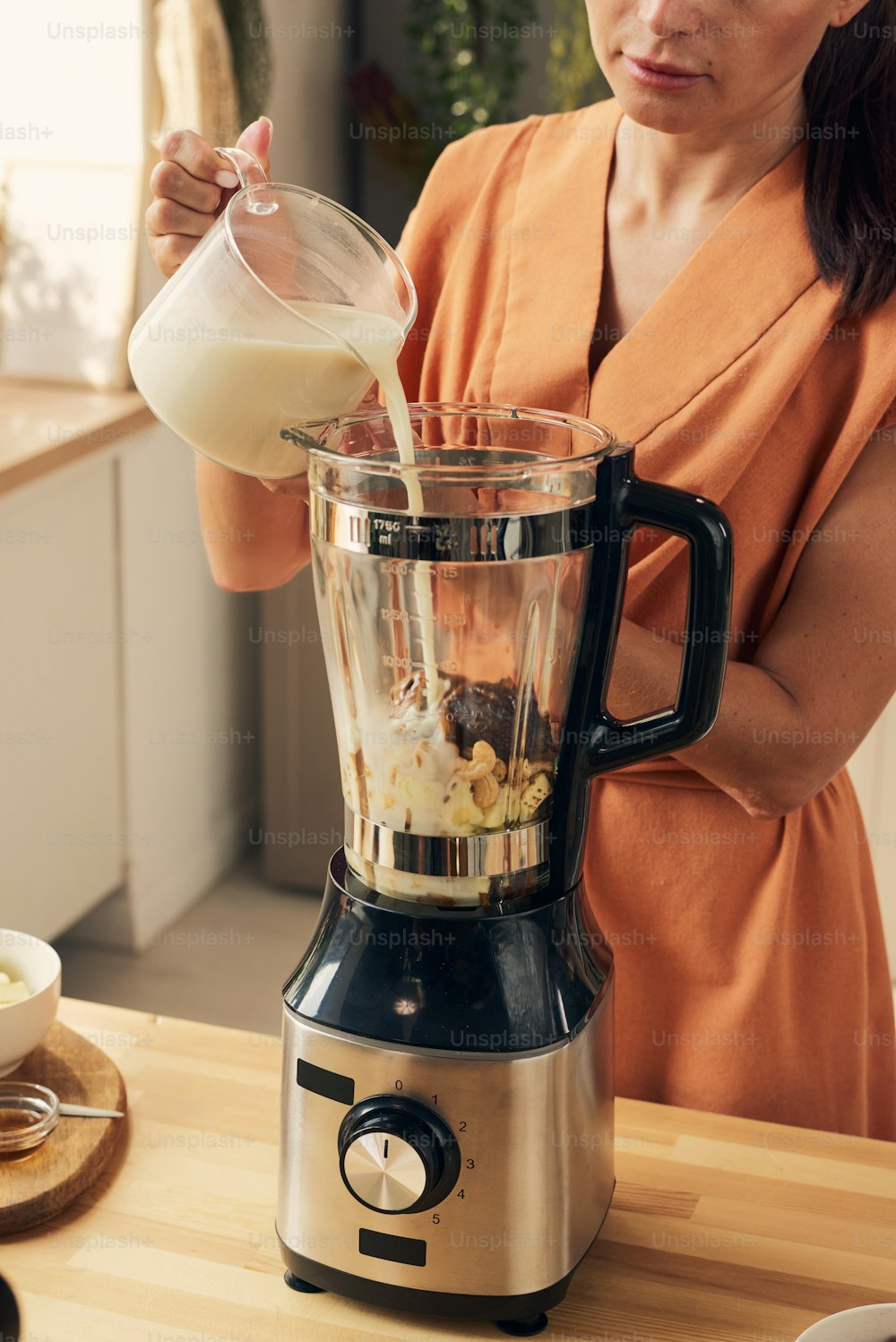Hausfrau im orangefarbenen Kleid gießt frische Milch mit gehackten Zutaten in den elektrischen Mixer, während sie Smoothie kocht