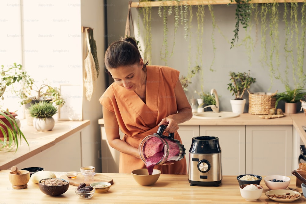 Jeune femme brune penchée sur la table de la cuisine tout en versant un smoothie aux légumes frais faits maison dans un bol