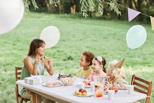 Portrait of young mother sitting at picnic table with group of kids during outdoor Birthday party in Summer, copy space