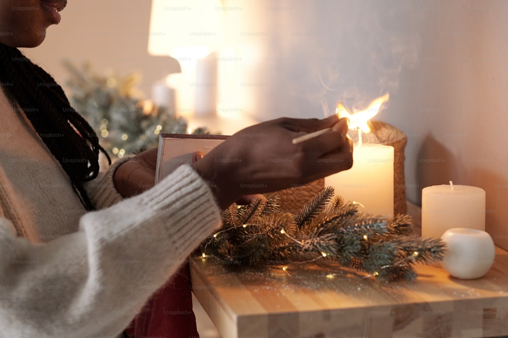 Hand of young female of African ethnicity lighting candles on wooden table decorated with coniferous branch before Christmas celebration
