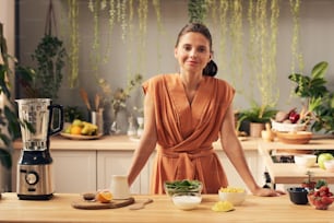 Mujer joven feliz mirándote mientras está de pie junto a la mesa de la cocina con frutas y verduras frescas antes de cocinar el batido
