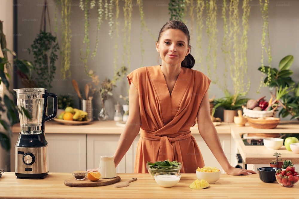 Mujer joven feliz mirándote mientras está de pie junto a la mesa de la cocina con frutas y verduras frescas antes de cocinar el batido