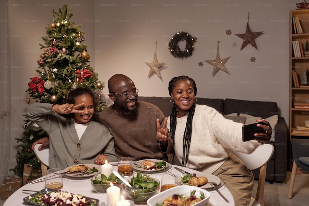 Feliz familia joven de tres haciendo selfies o comunicándose en video chat con sus amigos mientras están sentados junto a una mesa festiva