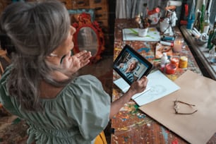 Visão de alto ângulo da mulher de cabelos grisalhos sentada à mesa e segurando o tablet enquanto olha para ele e conversa com seu bestie