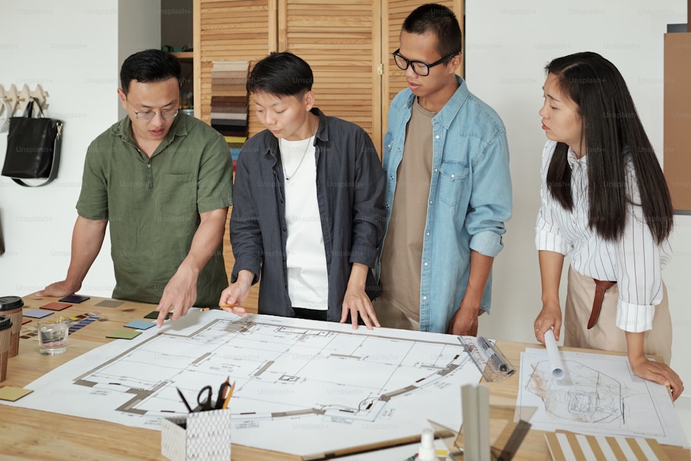 Group of young architects gathered by table with large sketch on blueprint discussing working points at meeting