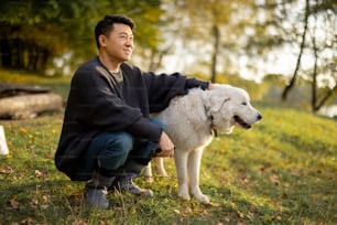 Satisfeito homem asiático sentado, acariciando seu Maremmano-Abruzzese Sheepdog e olhando para longe na natureza no dia ensolarado de outono. Conceito de descanso e fim de semana na natureza. Homem adulto usando botas e roupas quentes