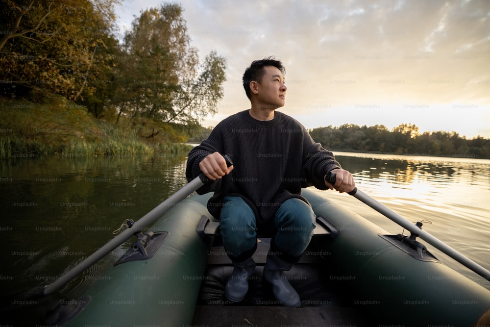 Asiatischer Mann, der am Herbstmorgen auf einem Schlauchboot im See oder Fluss schwimmt. Konzept der Erholung, des Wochenendes und des Urlaubs in der Natur. Idee der Freizeit im Freien. Erwachsene männliche Person, die Stiefel und warme Kleidung trägt