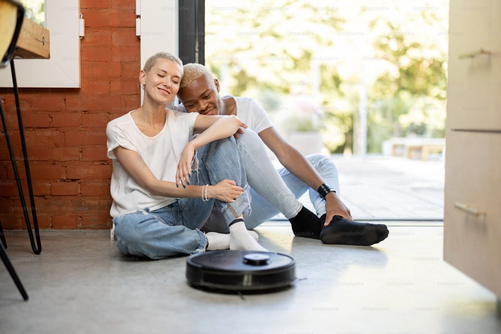 다인종 커플이 로봇 청소기로 집에 앉아 있다. 유럽 소녀와 흑인 남성이 함께 시간을 보낸다. 현대 가정 생활 방식의 개념입니다. 집 청소에 대한 아이디어