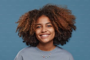 Retrato frontal de una joven afroamericana con cabello rizado natural sonriendo felizmente a la cámara en el estudio sobre fondo azul