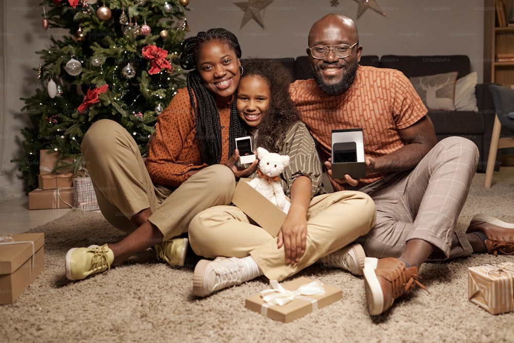 Miembros alegres de la familia con cajas de regalo abiertas que muestran sus regalos de Navidad mientras están sentados en el piso de la sala de estar