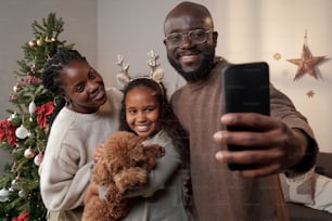 Des parents heureux et leur jolie fille avec un animal de compagnie regardant dans l’appareil photo du smartphone tout en faisant un selfie le jour de Noël