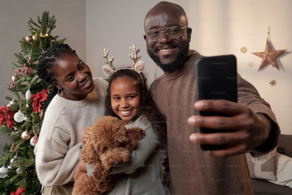 Padres felices y su linda hija con mascota mirando en la cámara del teléfono inteligente mientras se hacen selfie el día de Navidad