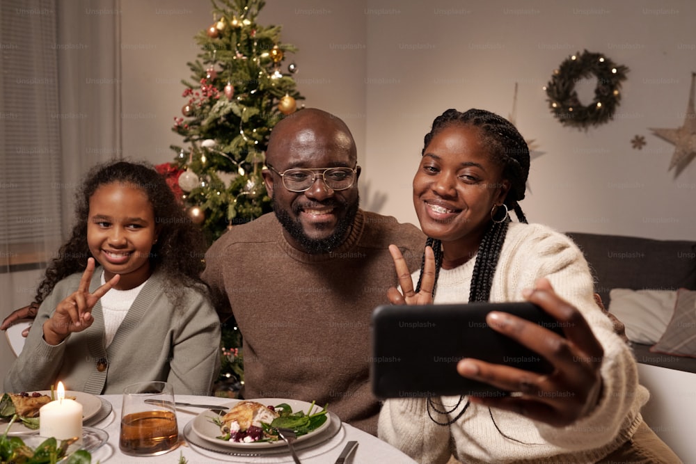 Three cheerful African people showing peace gesture while looking at smartphone camera during selfie by festive table on xmas day