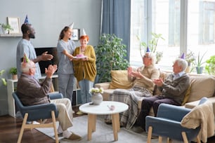 Nurse and doctor congratulating senior woman with her birthday and giving a cake to her with other senior people clapping hands