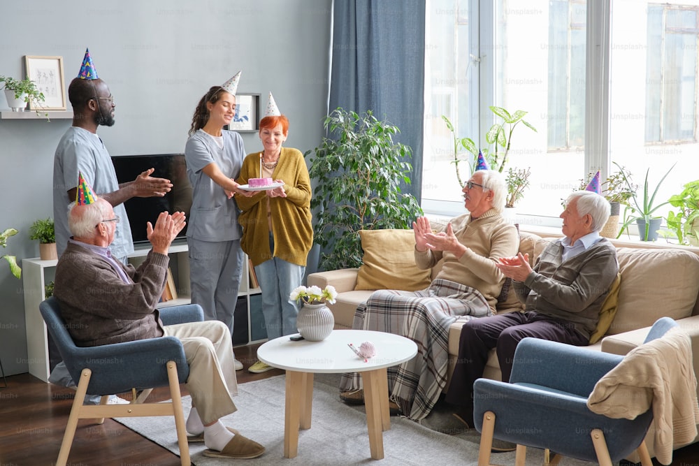Krankenschwester und Arzt gratulieren älterer Frau zu ihrem Geburtstag und geben ihr einen Kuchen mit anderen älteren Leuten, die in die Hände klatschen