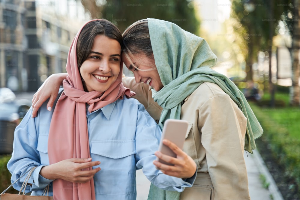 Nice selfie. Waist up of smiling young arabian woman wearing abayas taking self-portrait picture while bonding to each other. Happy millennial females making selfie on good quality smartphone camera