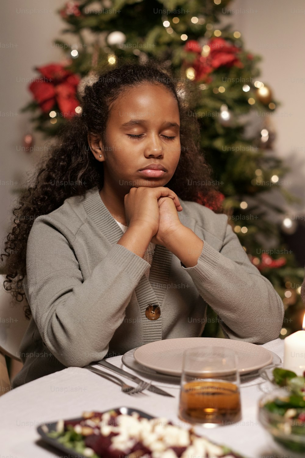 눈을 감고 턱으로 손을 모은 귀여운 혼혈 소녀가 축제 저녁 식사를 위해 준비된 테이블 옆에서 기도하고 있다