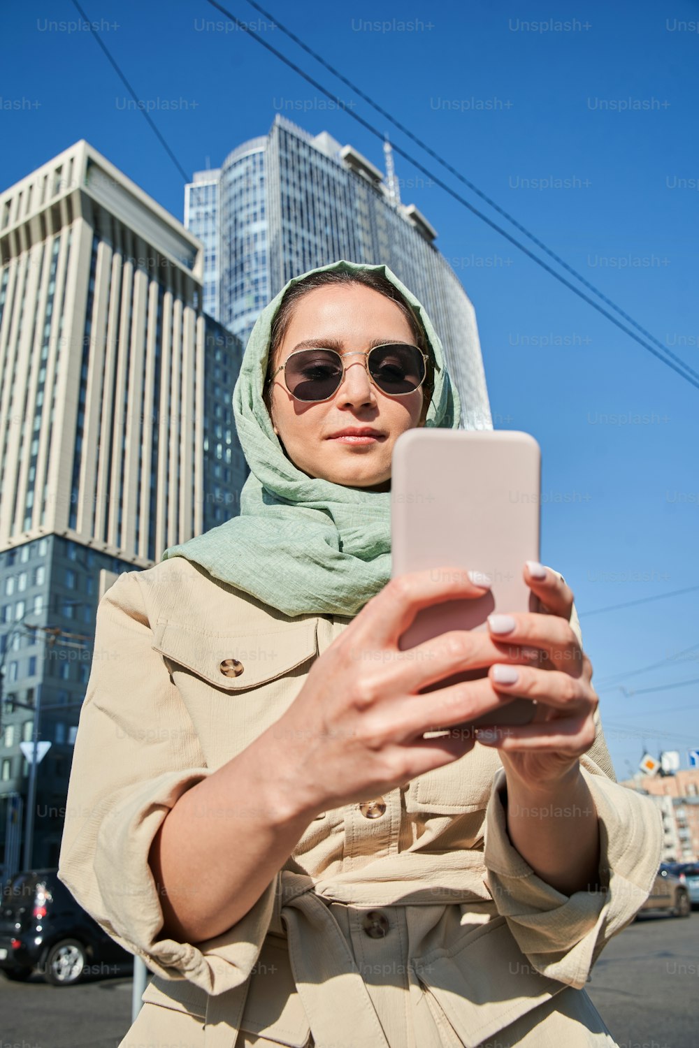 高い建物を背景にスマートフォンで自分撮りをする豪華なイスラム教徒の女の子のローアングルビュー。旅の間にカメラに向かってポーズをとる興奮した楽しい女性