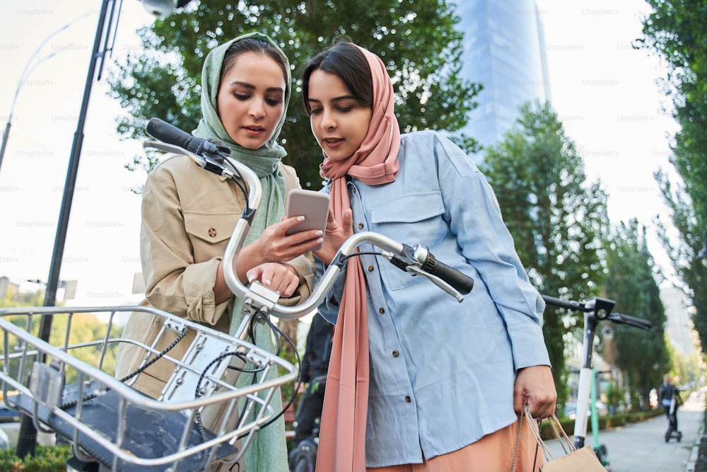 Summer day. Young millennial muslim woman in abayas using smartphone to unlocking urban bike with city street background. Bike rental concept