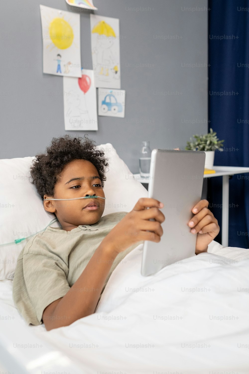 Petit garçon africain regardant quelque chose sur une tablette numérique allongé sur son lit dans une salle d’hôpital