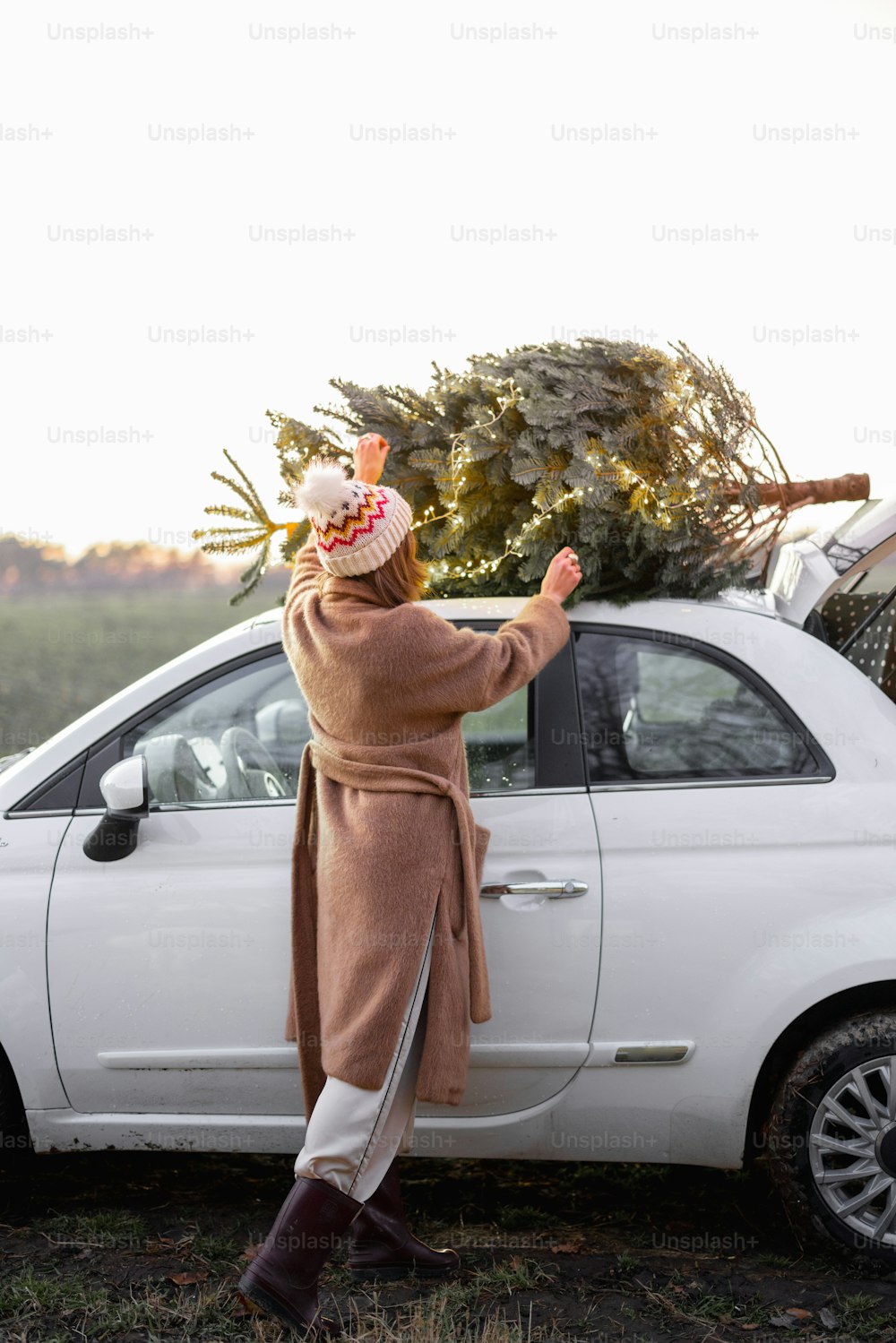 Frau packt Weihnachtsbaum auf einem Dach ihres Autos und bereitet sich auf einen Urlaub vor. Vorstellung von Weihnachtsstimmung und -feier. Frau trägt Pelzmantel und Hut. Abend mit schönem Sonnenuntergangshimmel