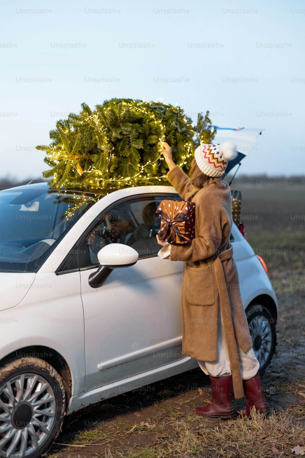 Donna che impacchetta l'albero di Natale su un tetto della sua auto, preparandosi per una vacanza. Idea di umore e celebrazione natalizia. Donna che indossa pelliccia e cappello. Serata con bellissimo cielo al tramonto