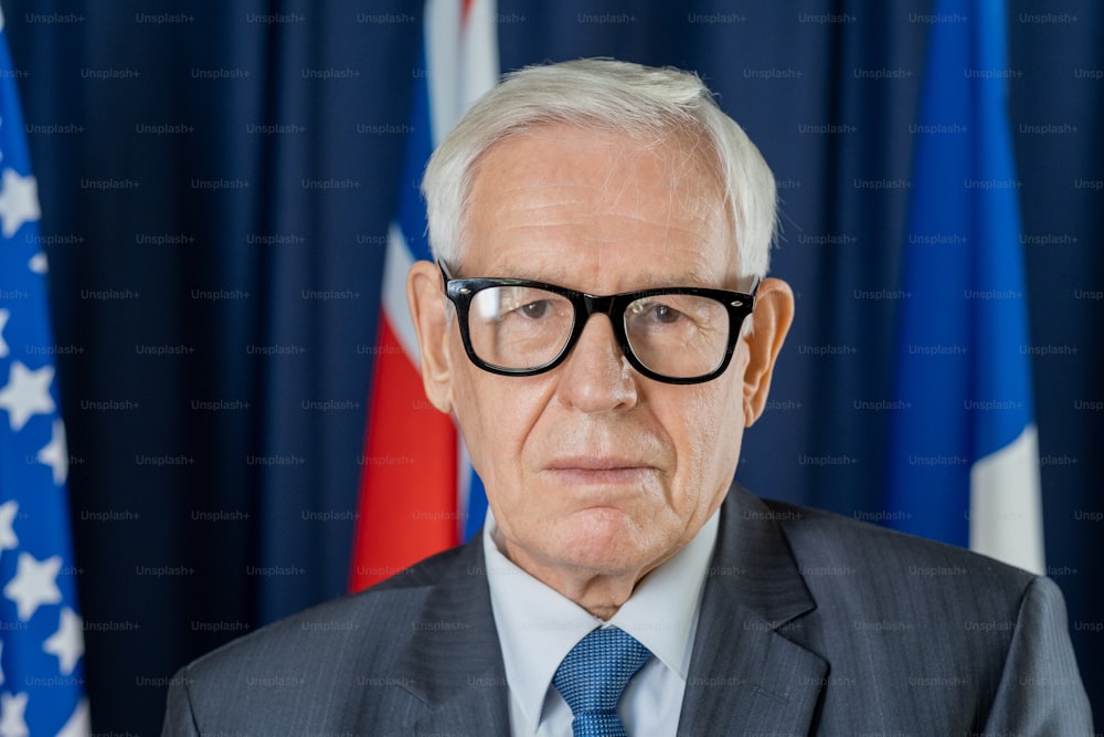 Portrait of serious confident senior male politician in eyeglasses standing against flags and dark blue curtains