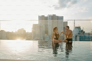 Glückliches junges Paar, das am Rande des Swimmingpools sitzt und mit Cocktailgläsern anstößt