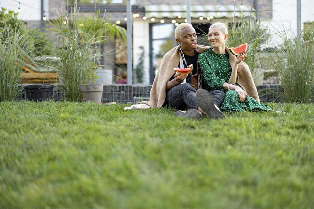 Multiracial couple eating watermelon in their garden. Concept of relationship and enjoying time together. Modern lifestyle. Black man hugging his european girlfriend. People covered in plaid