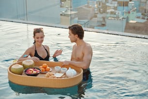 Lächelndes hübsches junges Paar, das das Frühstück im Pool auf dem Dach genießt