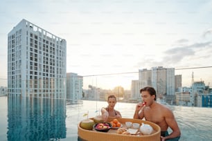 Attractive young man and woman eating delicious breakfast in rooftop swimming pool