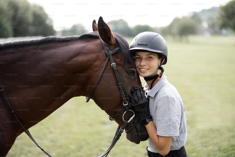 Female horseman hugging head of her brown Thoroughbred horse. Blurred image of green meadow in countryside. Concept of animal care. Rural rest and leisure. Idea of green tourism. Young european woman