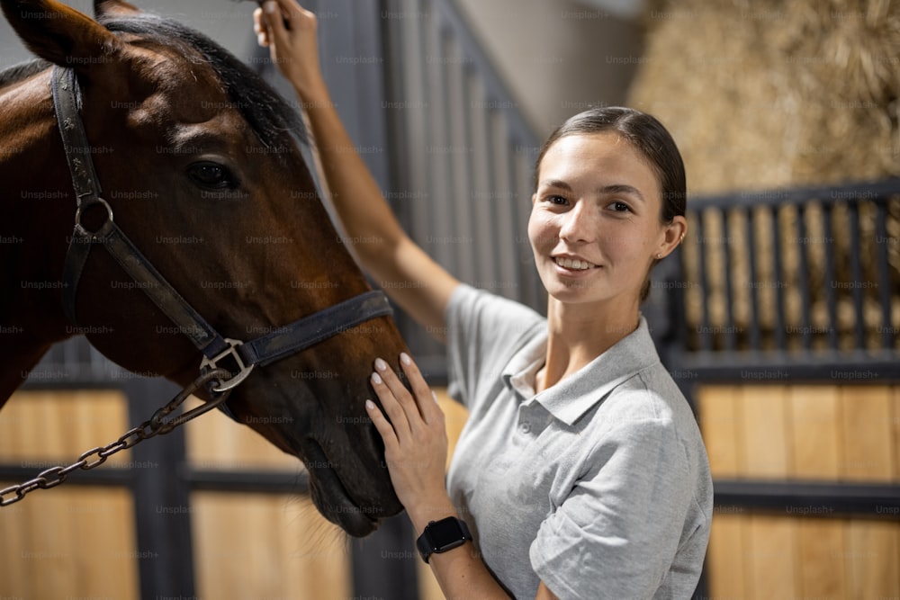 Portrait of female horseman with her brown Thoroughbred horse in stable. Concept of animal care. Rural rest and leisure. Idea of green tourism. Young european woman wearing uniform