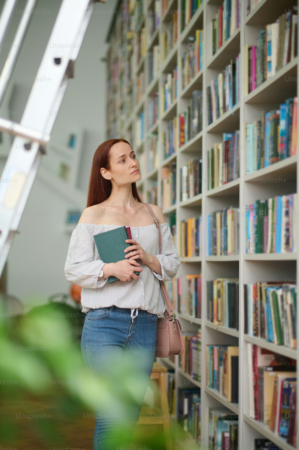 Selection of books. Young interested woman in blouse and jeans with handbag on shoulder choosing books near high bookshelves in library in afternoon