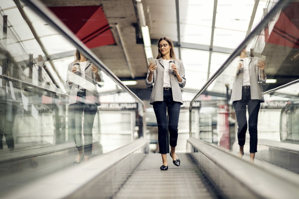 A busy, young businesswoman is going down the escalator with takeaway coffee and phone in her hands. A woman is replying on messages from her employees. A businesswoman on the escalator with a phone