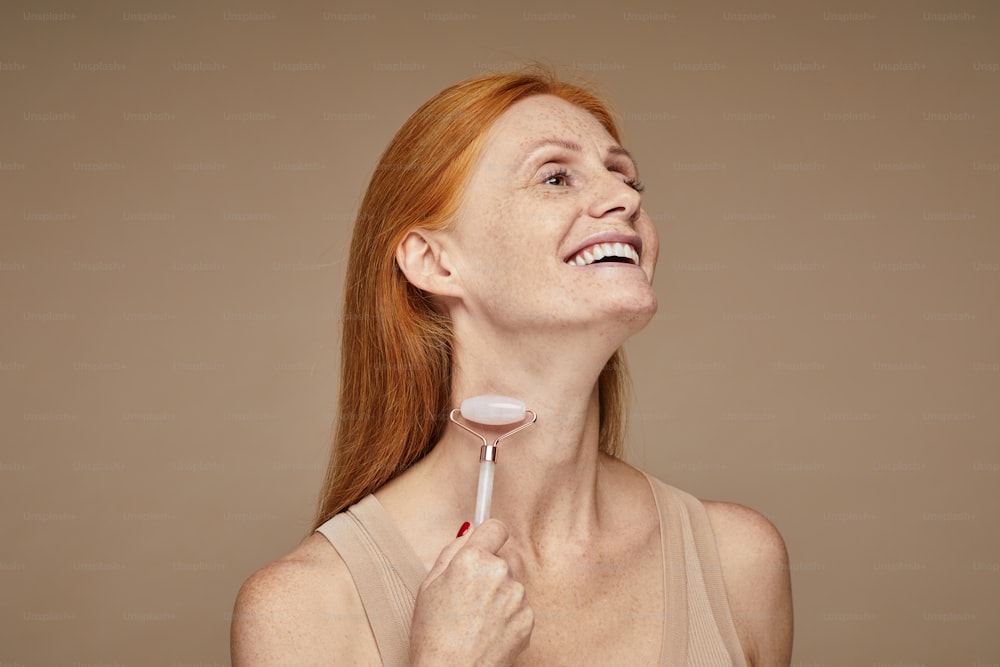 Portrait of beautiful red haired woman with freckles smiling and doing face massage using roller, copy space