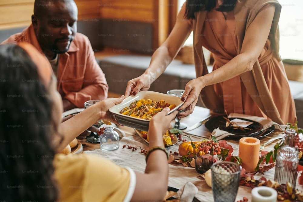Interracial girl helping her mother while taking bowl with homemade food over served festive table during family dinner