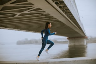 Hübsche junge Frau im blauen Trainingsanzug, die am Herbstmorgen am Fluss entlang läuft