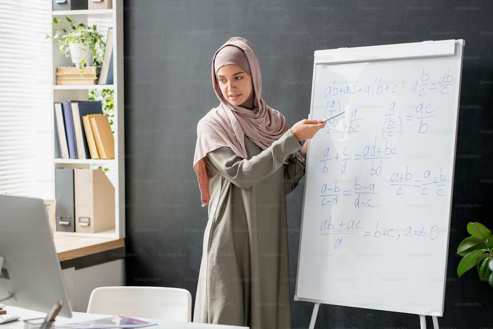 Confident teacher of algebra pointing at formula or equation on whiteboard while explaining it to students during online lesson