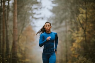 Young woman in blue track suit running toward camera on the forest trail at autumn