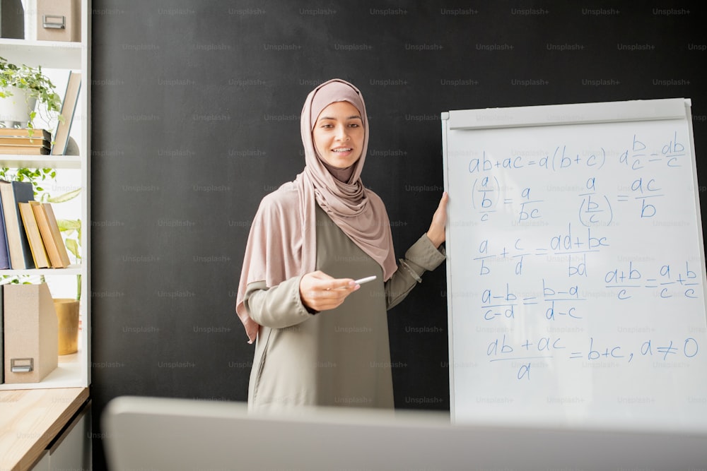 Happy young teacher in hijab standing by whiteboard with algebraic formula and equations while explaining them in front of computer