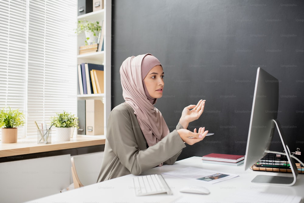 Jeune enseignante musulmane avec un stylo expliquant quelque chose au public en ligne tout en étant assise devant un écran d’ordinateur