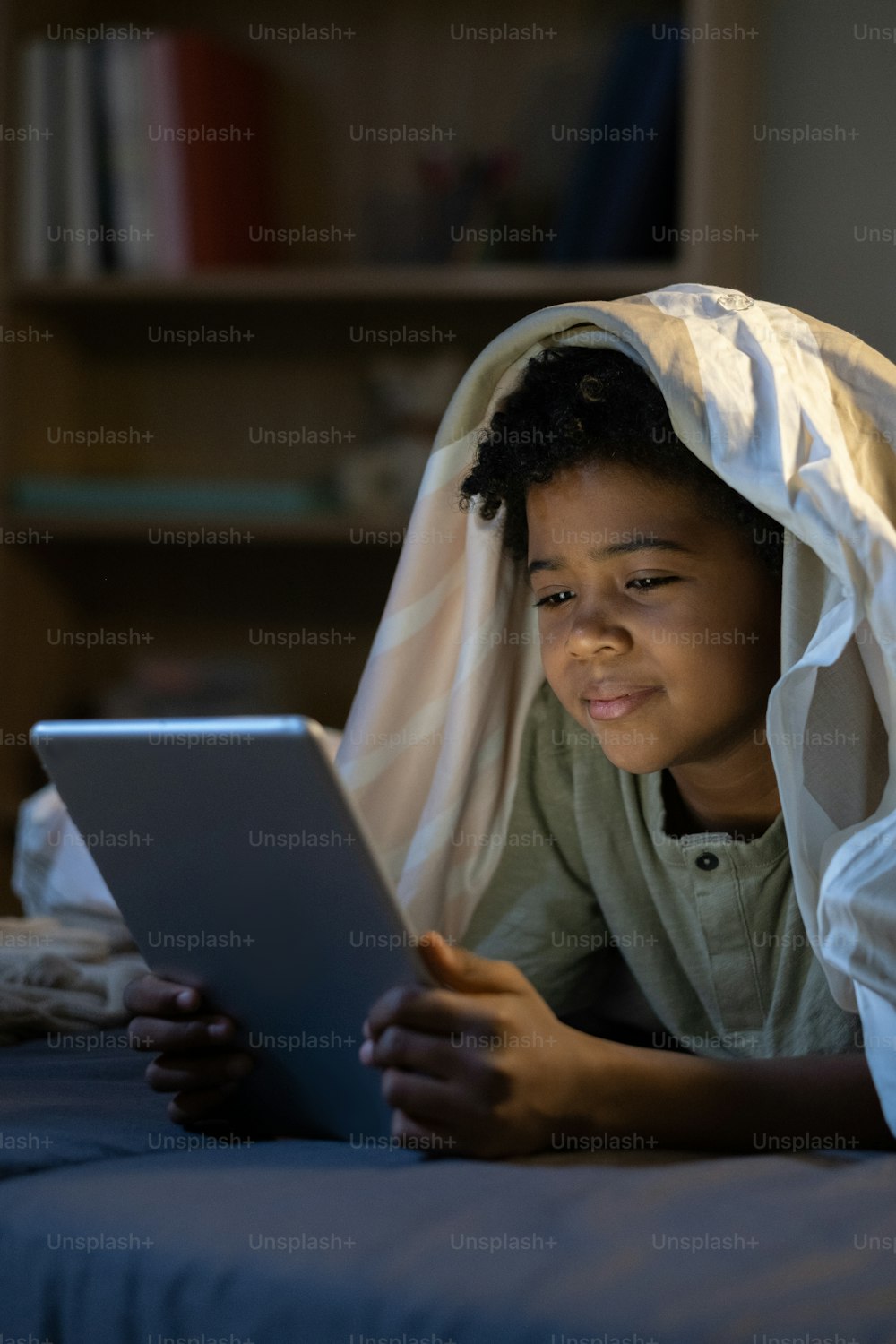 Content African American boy lying with tablet under duvet in dark nursery room
