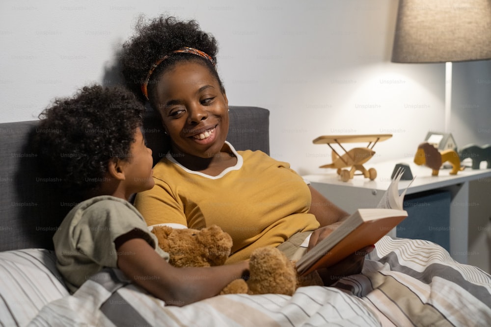 Giovane madre afroamericana allegra seduta sotto il piumone nel letto e leggendo fiabe al figlio