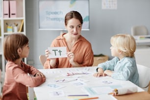 Jeune professeur d’anglais montrant la carte avec lettre aux enfants et ils l’apprennent à table pendant une leçon